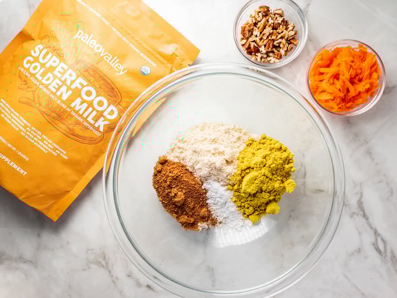Baking ingredients for healthy carrot cake muffins, featuring Paleovalley Superfood Golden Milk, shredded carrots, and chopped pecans.