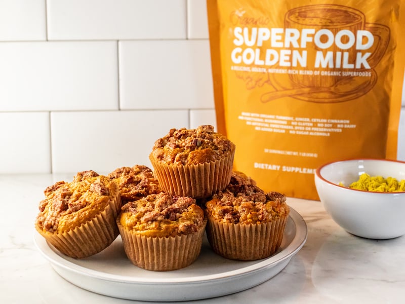 Freshly baked healthy carrot cake muffins made with Paleovalley Superfood Golden Milk, displayed on a white plate with the supplement pouch and a bowl of golden powder in the background.