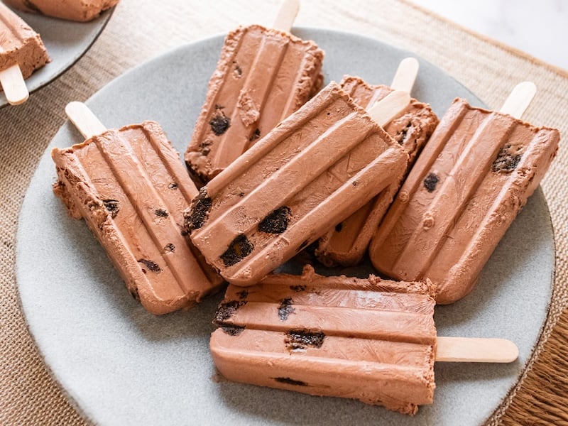 Creamy chocolate fudge popsicles with chunks of dark superfood bars throughout, stacked on a soft gray plate, showcasing a rich and indulgent frozen paleo treat.