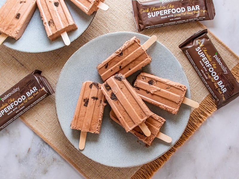 Chocolate fudge popsicles with chunks of Paleovalley Dark Chocolate Chip Superfood Bar on a gray ceramic plate, surrounded by additional bars and popsicles on a burlap placemat and marble surface.