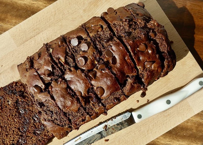 Sliced chocolate banana bread with chocolate chips and chocolate bone broth powder on a wooden cutting board, next to a serrated knife.