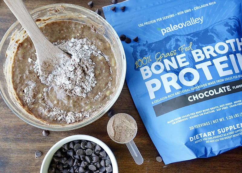 Bowl of chocolate banana bread batter with Paleovalley Chocolate Bone Broth Protein being mixed in, surrounded by chocolate chips and a protein scoop.