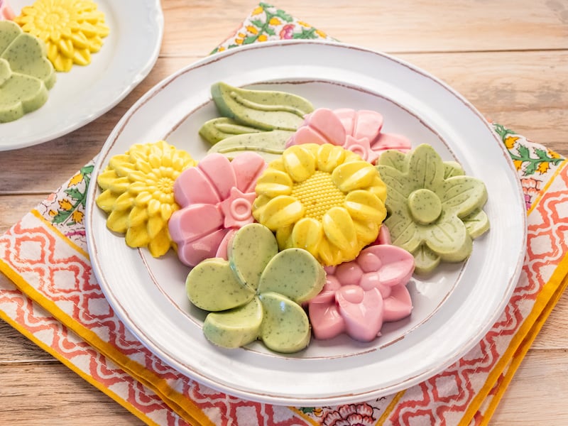 A plate of creamy electrolyte gummies in pastel shades of yellow, pink, and green, molded into playful floral and leaf shapes, displayed on a colorful napkin with a wooden table backdrop.