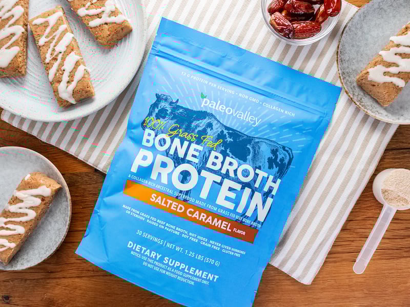 Homemade paleo protein bars drizzled with icing, made using Paleovalley Salted Caramel Bone Broth Protein, displayed with a scoop of powder and a pouch on striped linens and ceramic plates.
