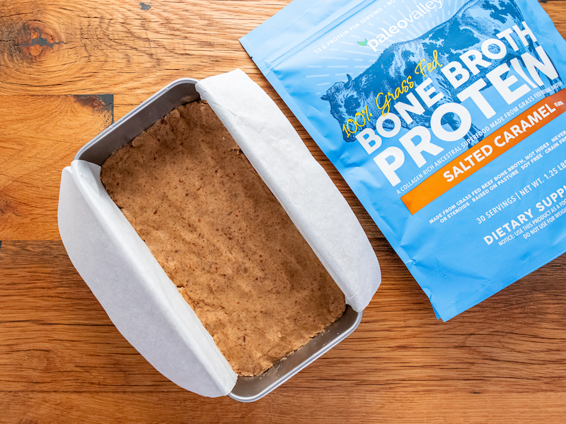 Homemade paleo protein bar mixture pressed into a parchment-lined loaf pan, made using Paleovalley Salted Caramel Bone Broth Protein, shown beside the product bag on a wooden surface.
