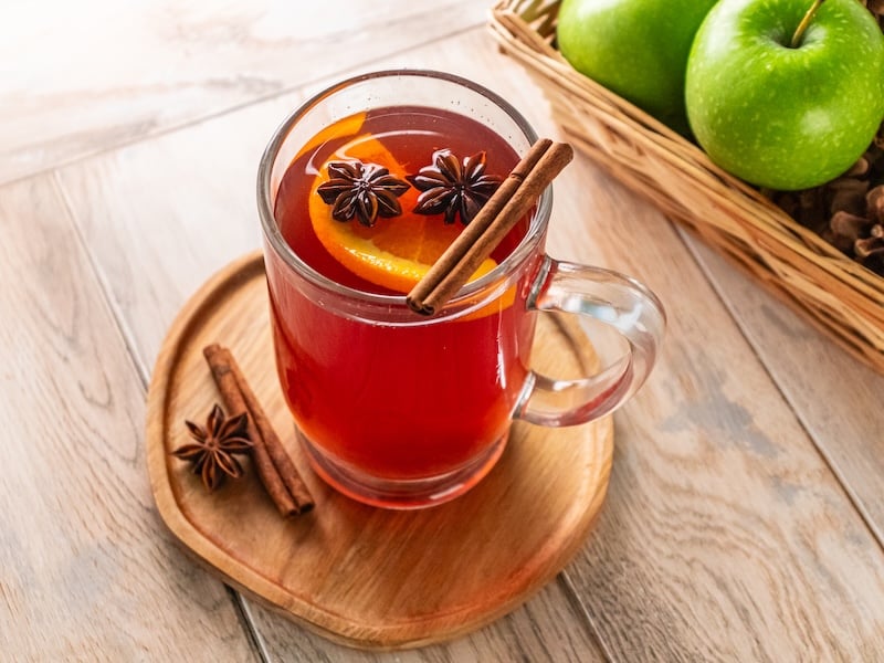 Warm elevated hot apple cider garnished with cinnamon sticks, star anise, and orange slices, served in a clear mug on a wooden tray.