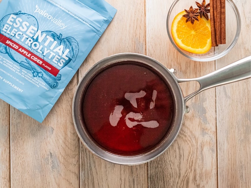 Hot apple cider simmering in a saucepan made with Paleovalley Essential Electrolytes Spiced Apple Cider, with orange slices and cinnamon nearby.
