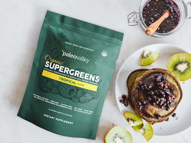 Paleovalley Organic Supergreens Tropical flavor pouch on a marble surface with green pancakes topped with wild blueberries and kiwi slices.