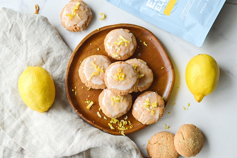 Glazed lemon cookies topped with zest, made with Paleovalley Essential Electrolytes, displayed on a wooden plate with fresh lemons.