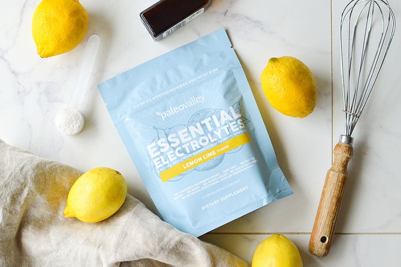 Paleovalley Essential Electrolytes Lemon Lime flavor surrounded by fresh lemons, a whisk, and baking ingredients on a marble counter.