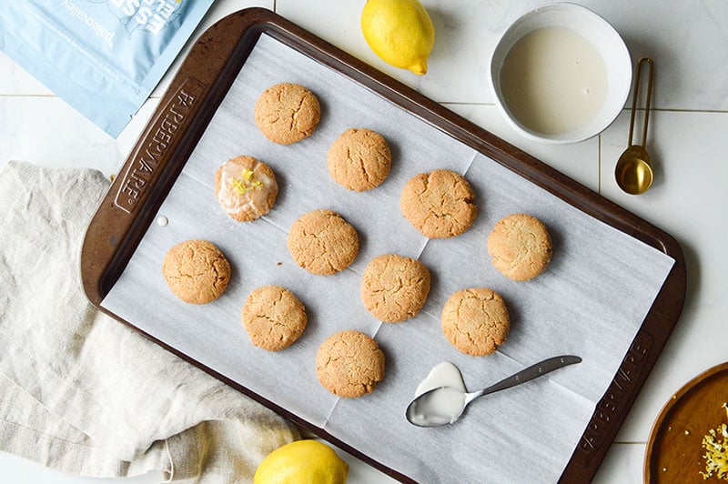 Freshly baked lemon cookies on a parchment-lined baking tray, with one cookie topped with glaze and lemon zest, surrounded by baking tools and ingredients.