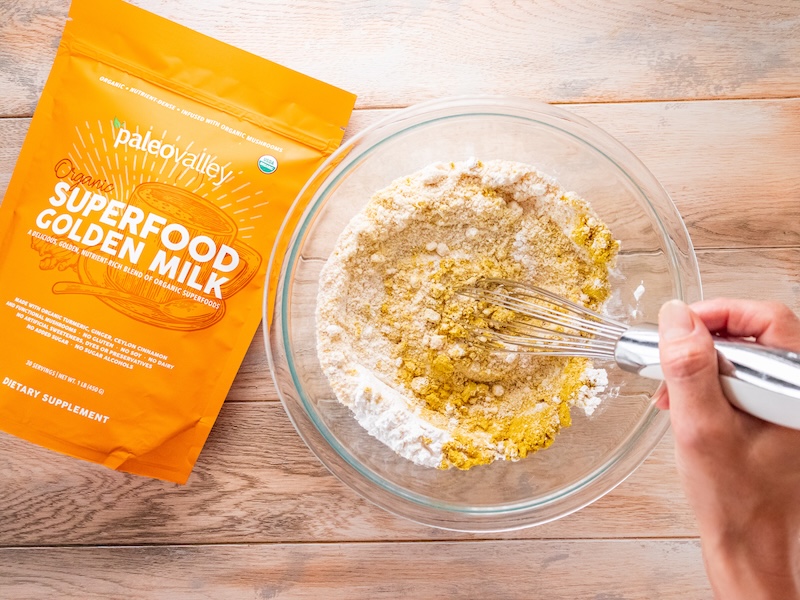 Hand whisking together coconut flour and Paleovalley Superfood Golden Milk powder in a glass bowl, next to a bright orange bag of the supplement.