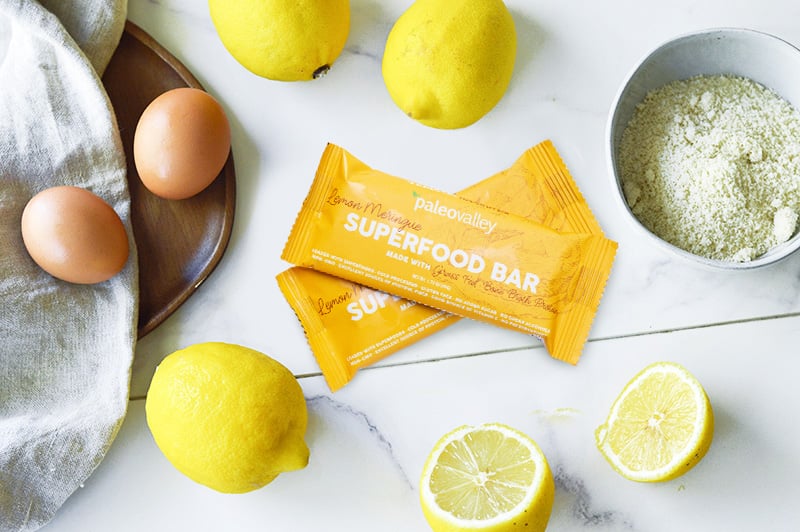 Paleovalley Lemon Meringue Superfood Bars surrounded by fresh lemons, eggs, and almond flour on a white marble surface.