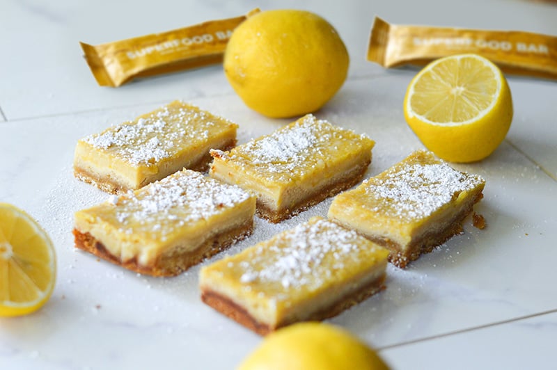 Paleo lemon coconut bars made with Paleovalley Lemon Meringue Superfood Bar, dusted with powdered sugar and surrounded by fresh lemons.