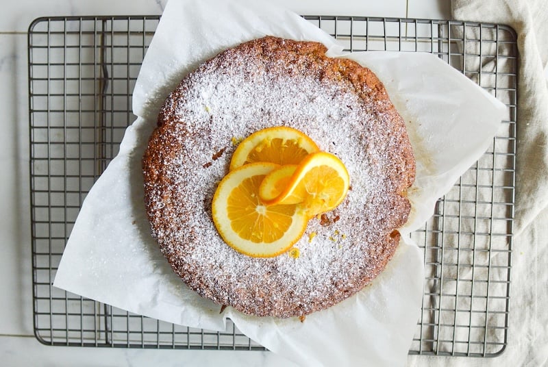 Orange almond cake topped with powdered sugar and fresh orange slices, cooling on a wire rack with parchment paper.