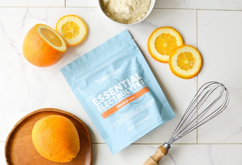 Paleovalley Essential Electrolytes in Orange flavor on a tiled surface, surrounded by fresh orange slices, a whisk, and baking ingredients.