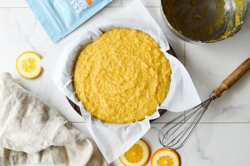 Orange almond cake batter in a parchment-lined baking pan, surrounded by sliced oranges, a whisk, a mixing bowl, and Paleovalley Essential Electrolytes.