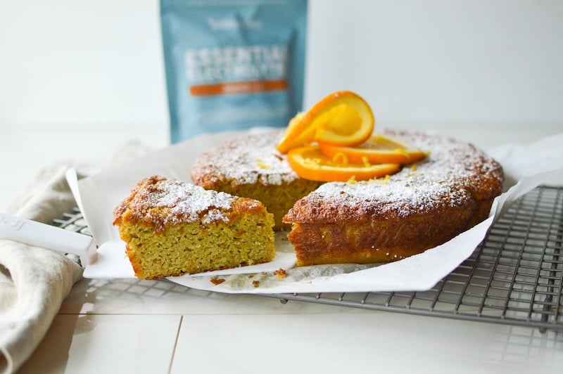 Sliced orange almond cake topped with powdered sugar and orange slices, served on parchment paper with Paleovalley Essential Electrolytes in the background.