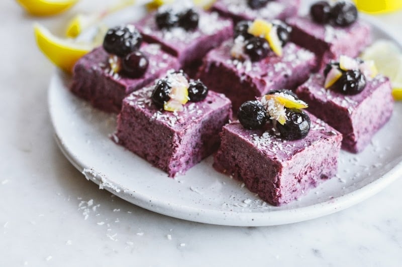 Plate of blueberry dessert bars topped with blueberries, lemon zest, and shredded coconut on a marble surface.
