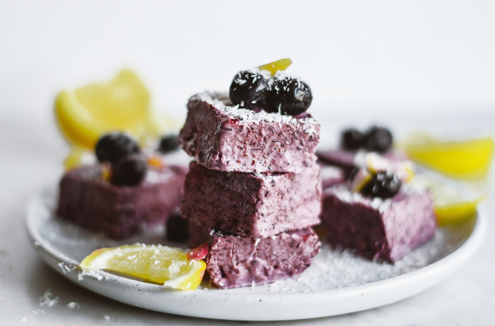 Stack of blueberry dessert bars topped with blueberries, lemon zest, and shredded coconut on a white plate with lemon wedges.