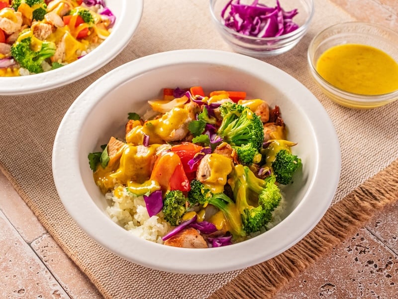 Bowl of paleo Thai chicken over cauliflower rice, topped with roasted broccoli, bell peppers, purple cabbage, and drizzled with golden turmeric sauce, served with extra sauce and shredded cabbage on the side.