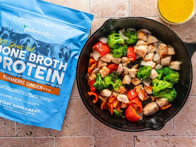 Colorful chicken and vegetable stir-fry with broccoli, red bell peppers, and onions in a cast iron skillet, next to a packet of Paleovalley 100% Grass Fed Bone Broth Protein in Turmeric Ginger flavor and a small bowl of golden sauce.