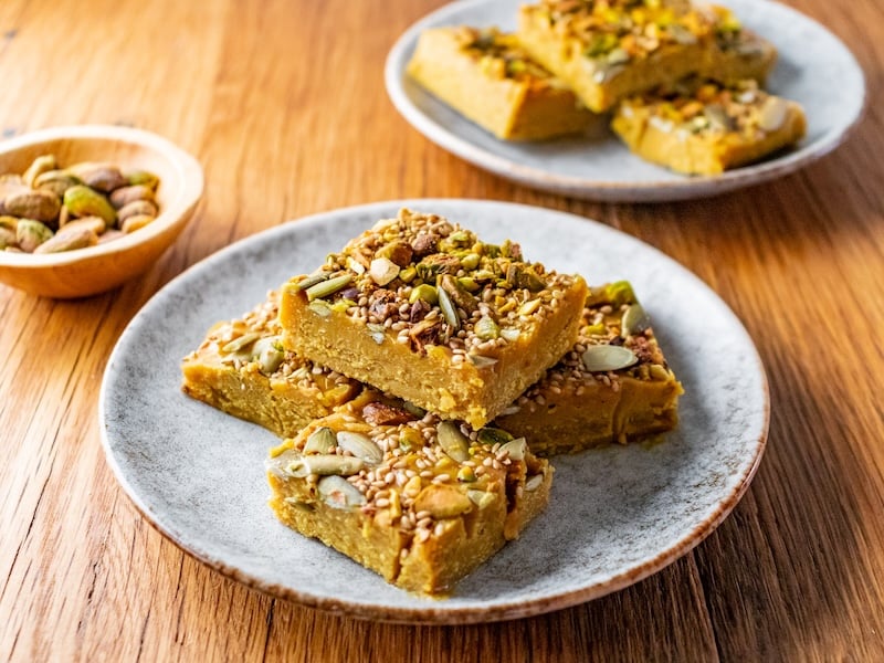Golden superfood fudge squares stacked on a ceramic plate, topped with sesame seeds, pistachios, and pumpkin seeds, with a bowl of pistachios and extra fudge in the background.