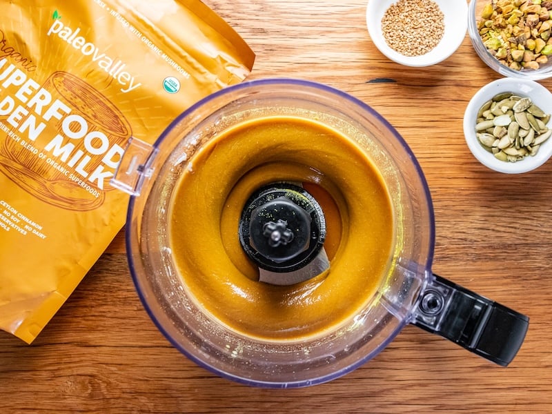 Creamy golden fudge mixture blending in a food processor, with Paleovalley Superfood Golden Milk bag and bowls of seeds and chopped nuts in the background on a wooden surface.