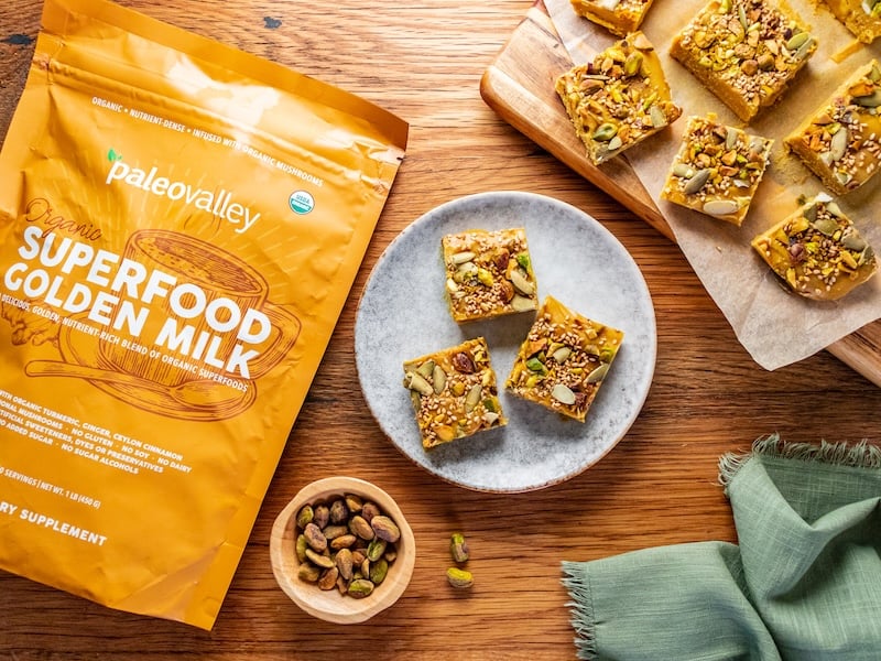 Overhead view of golden superfood fudge squares on a ceramic plate and parchment-covered board, surrounded by a Paleovalley Superfood Golden Milk bag, pistachios, and a green napkin on a wooden surface.