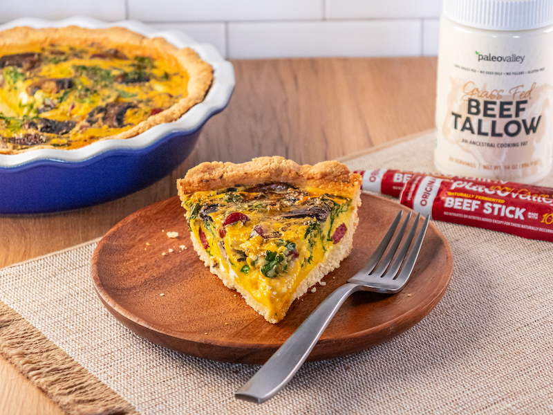 A slice of tallow-crusted quiche with mushrooms, spinach, and Paleovalley beef sticks served on a wooden plate with a fork.