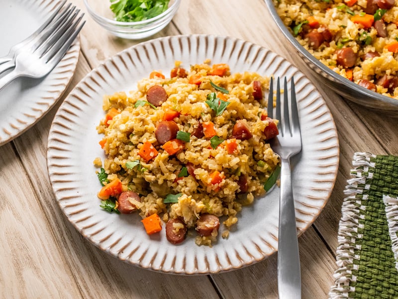Cauliflower fried rice with chopped veggies and sliced teriyaki chicken sticks, served on a ceramic plate with a fork.