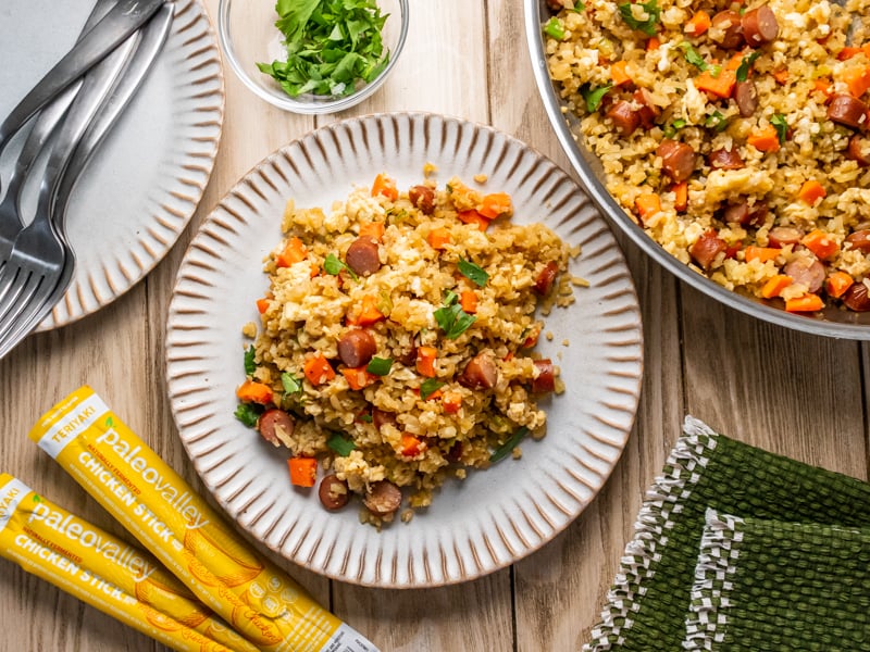 Cauliflower fried rice with diced carrots, vegetables, and sliced teriyaki chicken sticks, garnished with fresh parsley in a stainless steel skillet.