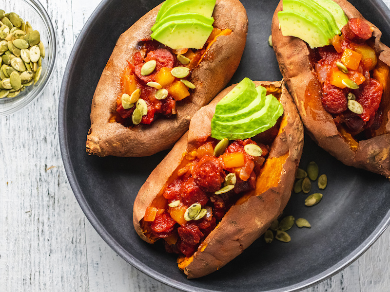 Three baked sweet potatoes topped with venison chili, avocado, and pumpkin seeds on a dark plate with PaleoValley venison sticks nearby.