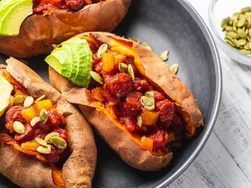 Close-up of baked sweet potatoes stuffed with venison chili, topped with avocado slices and pumpkin seeds, served on a dark plate.