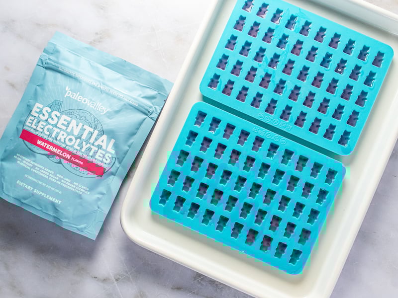 Homemade watermelon electrolyte gummies made with Paleovalley Essential Electrolytes, poured into bright blue gummy bear molds and set on a white tray, with the product pouch beside them on a marble surface.