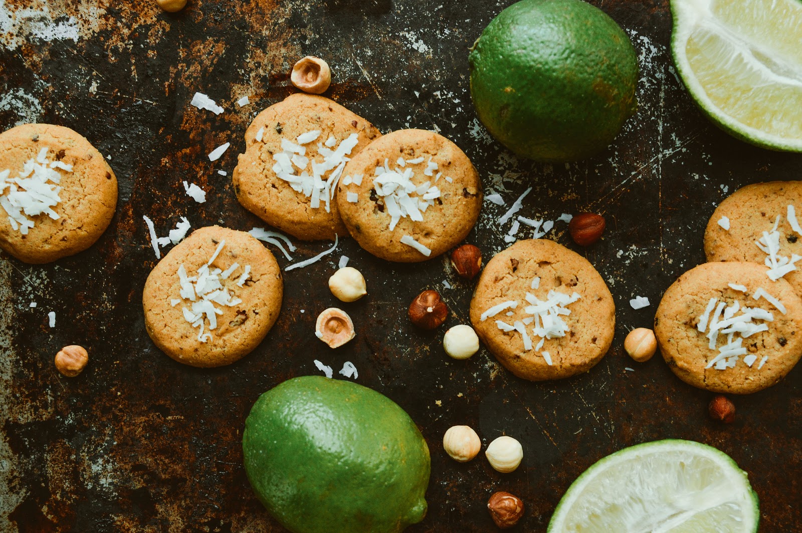 Golden brown paleo lime cookies topped with shredded coconut, scattered with hazelnuts and whole limes on a rustic baking sheet, highlighting tropical flavor and texture.