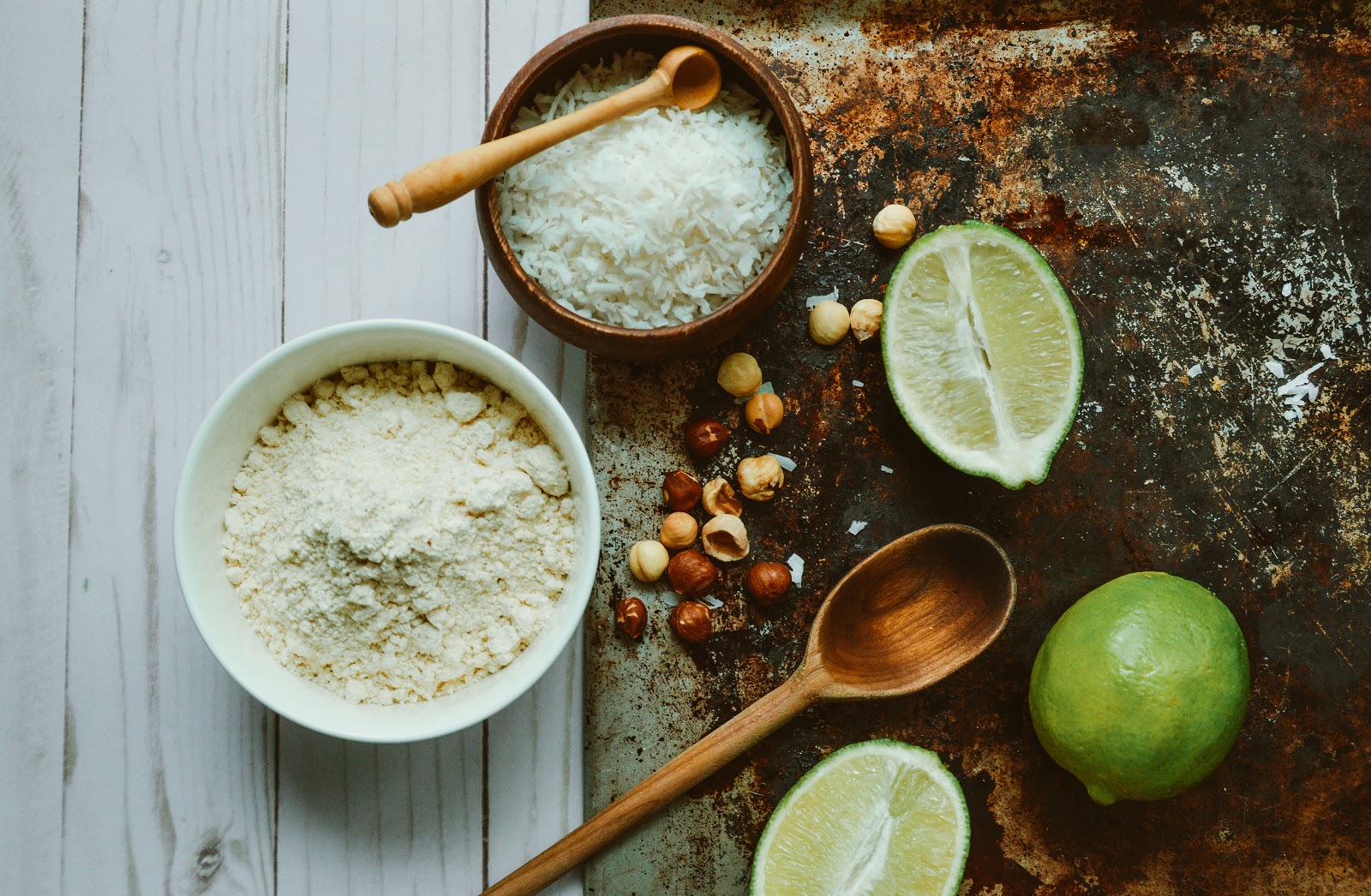 Flat lay of cookie ingredients including almond flour, shredded coconut, chopped hazelnuts, fresh limes, and wooden utensils on a rustic surface, styled for tropical-inspired paleo baking.