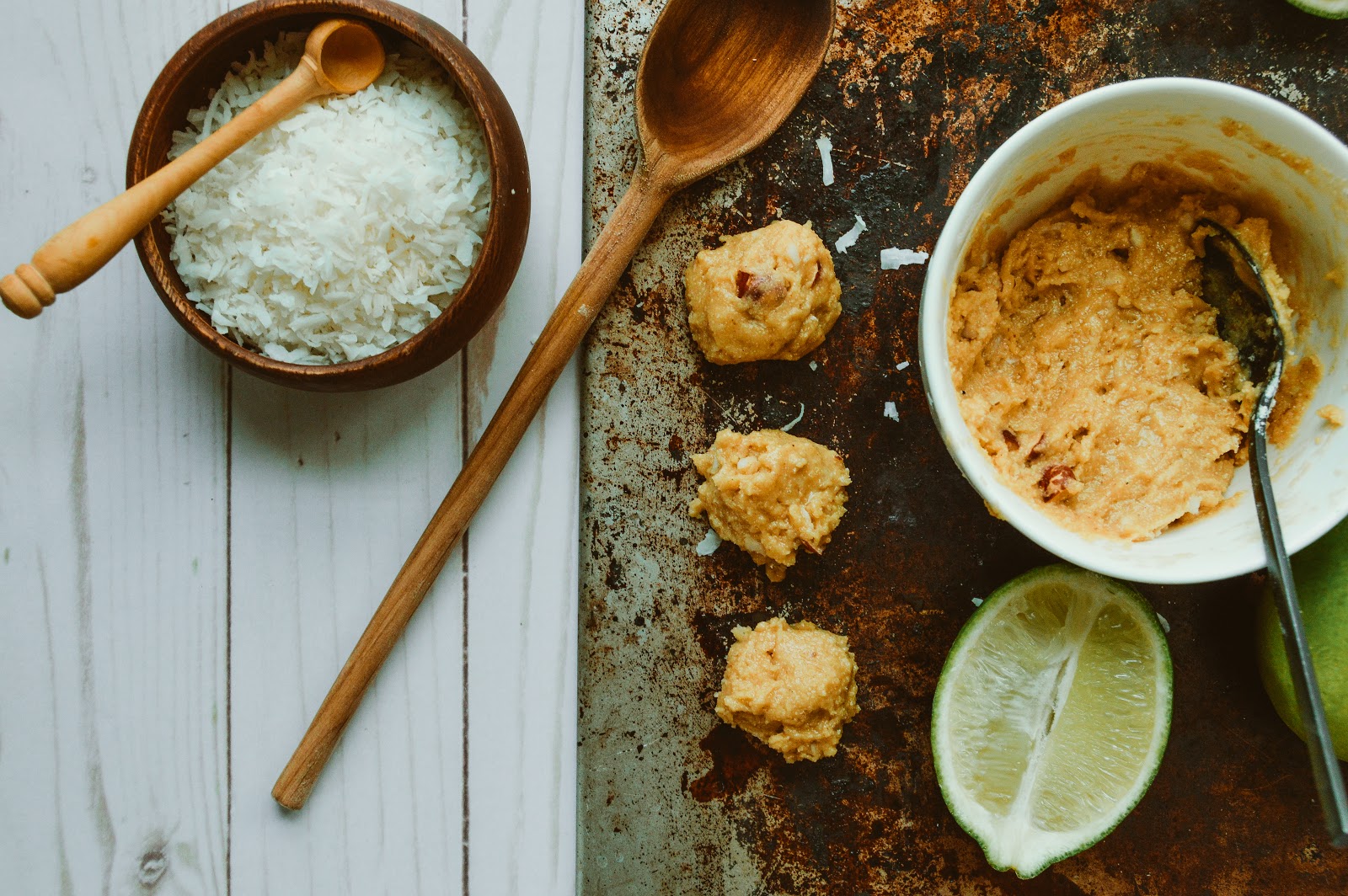 Paleo protein bar dough in a white mixing bowl with spoon, portioned scoops arranged on a rustic baking sheet, surrounded by shredded coconut, lime wedges, and wooden utensils.