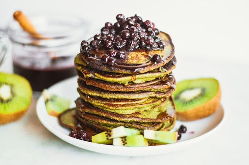 Stack of green pancakes topped with wild blueberries and syrup, served with sliced kiwi on a white plate.