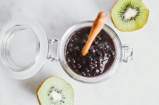 Jar of wild blueberry sauce with a wooden spoon, surrounded by sliced kiwi on a marble surface.