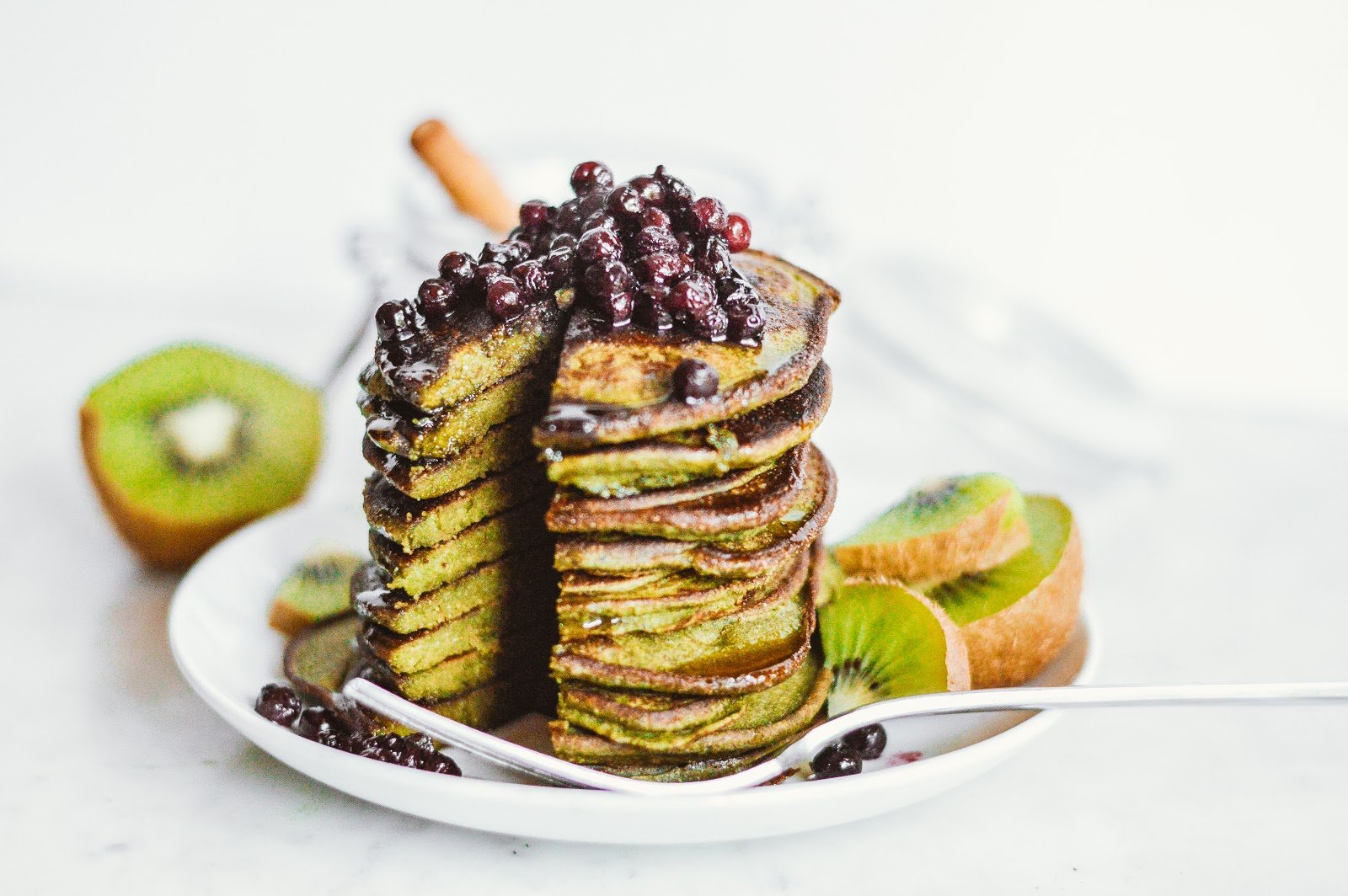 Stack of green pancakes with a cut section, topped with wild blueberries and syrup, served with sliced kiwi on a white plate.